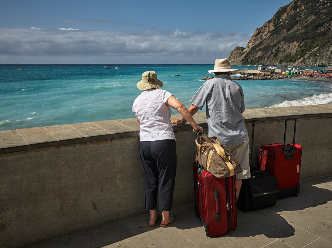 Two travelers look towards the ocean with their luggage.